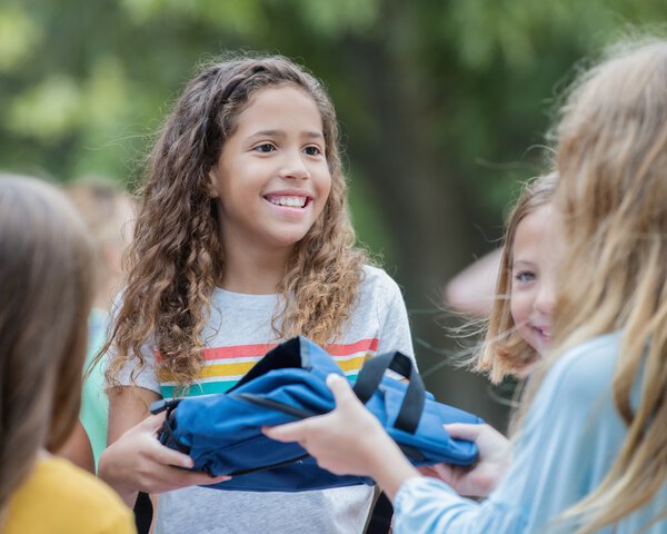 Lächelndes junges Mädchen übergibt anderem Mädchen einen Rucksack | © Courtney Hale - Getty Images