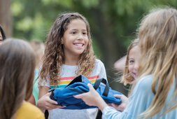 Lächelndes junges Mädchen übergibt anderem Mädchen einen Rucksack | © Courtney Hale - Getty Images